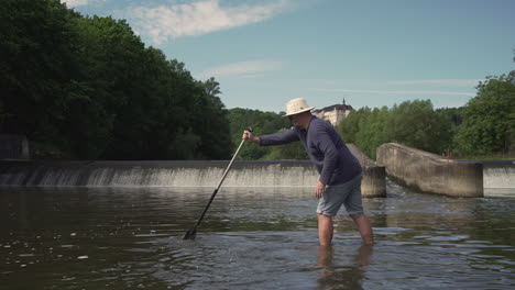 Male-paddler-checking-water-level-of-Sazava-river-with-paddle,-weir-in-background