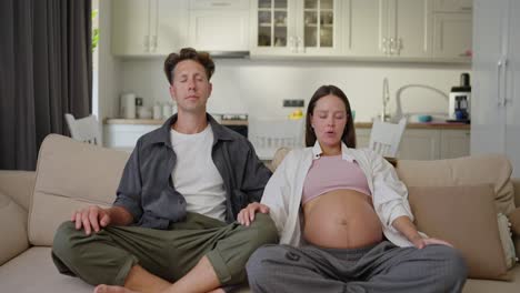 confident middle aged man along with his pregnant wife sit on the sofa and meditate during their leisure time at home on the sofa while waiting for the baby at home