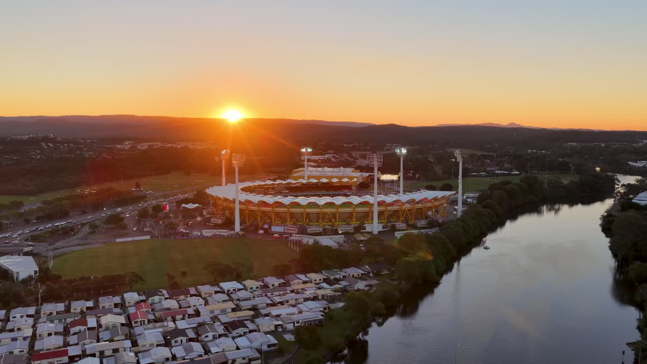 Drone captures a sweeping aerial view of a stadium by the river at sunset, with glowing stadium lights, calm water, and distant hills
