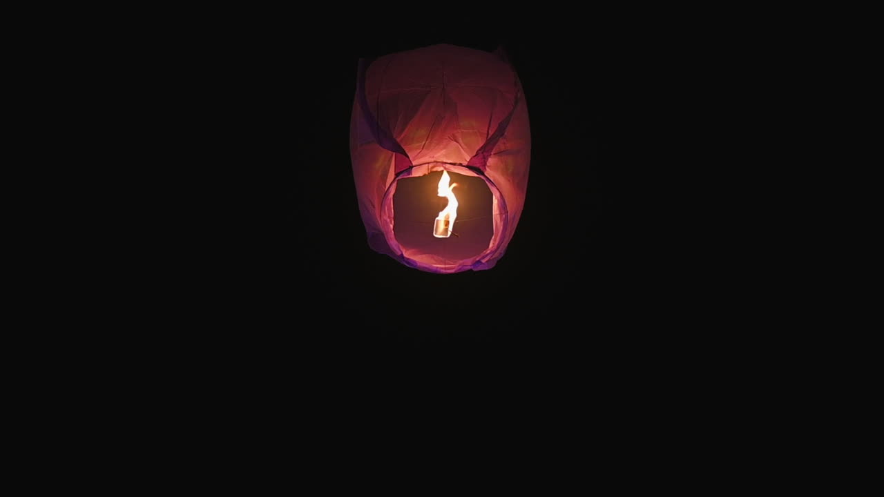 A single glowing lantern floats upward in the dark night sky, filmed in slow motion