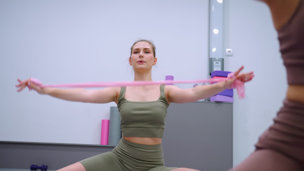 primer plano de una mujer de marrón participando en un ejercicio de fitness, estirándose con una cinta en un gimnasio, junto a otra mujer de verde, con equipos dispuestos en una mesa