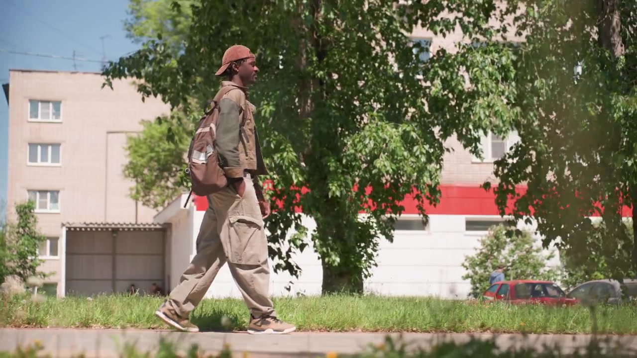 Close up of man walking along grassy path with cars and buildings in background. Man carries brown backpack while wearing casual outdoor clothing including cargo pants and cap. scene