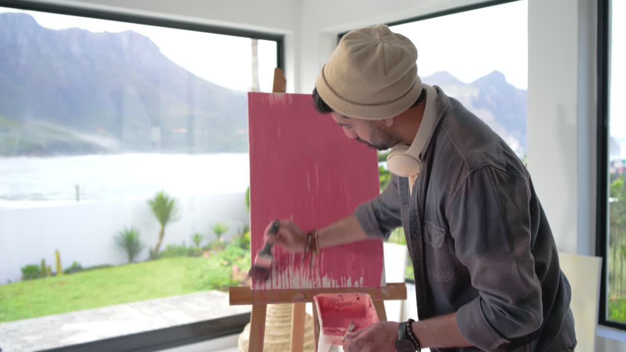 Indian man painting at easel, dipping brush in pink tray and creating dripping canvas wash
