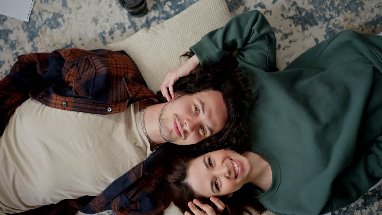 Camera rotation portrait of a happy brunette guy with curly hair lies head to head together with a brunette girl on a pillow on the floor at home in a modern apartment
