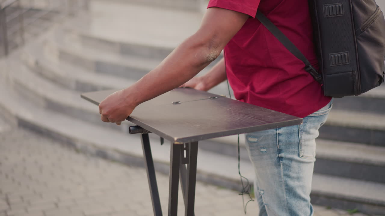 black street musician sets up turntable on folding table near stone steps, wearing red shirt and jeans, backpack visible, hands adjusting deck and controls, urban daytime busking vibe, focused