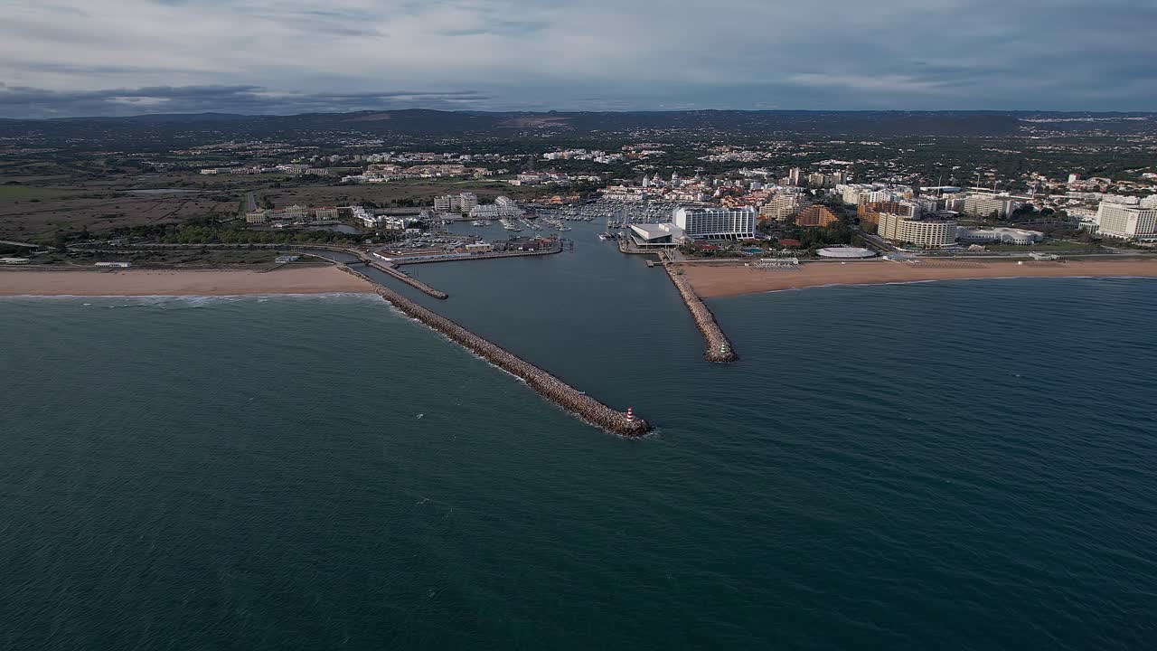 Drone approach from Atlantic Ocean towards Algarve coastline framing twin breakwaters and red‑and‑white Farol de Vilamoura lighthouse at channel entrance to Marina de Vilamoura in Quarteira, Portugal