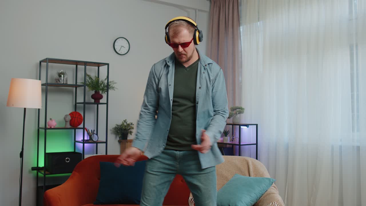 Overjoyed young man in wireless headphones dancing singing on cozy couch in living room at home