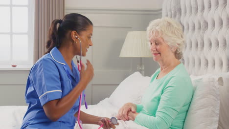 female care worker in uniform listening to chest of senior woman at home in bed with stethoscope