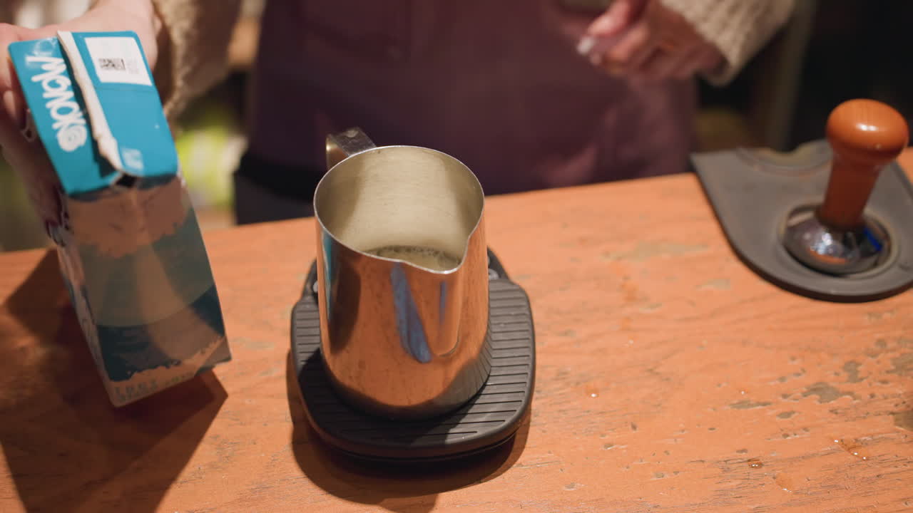 Close up of bartender pouring milk from carton into stainless steel frothing cup placed on scale, with tamper nearby on wooden counter in warm cafe setting during drink preparation process