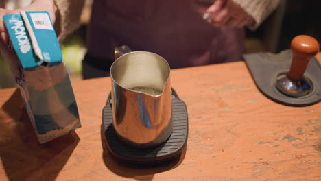 close up of bartender pouring milk from carton into stainless steel frothing cup placed on scale, with tamper nearby on wooden counter in warm cafe setting during drink preparation process