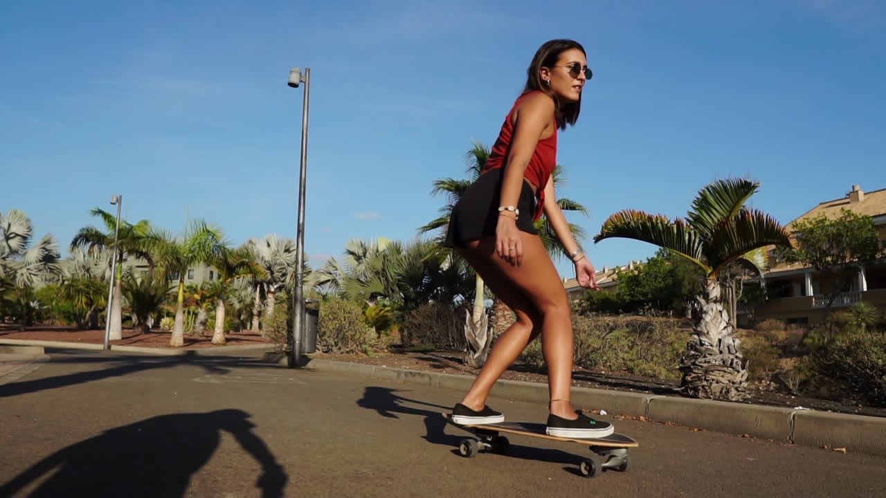 A young woman, shorts and sneakers completing her summer look, rides a longboard by palm trees in slow motion