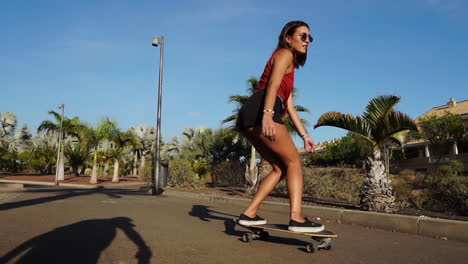 a young woman, shorts and sneakers completing her summer look, rides a longboard by palm trees in slow motion