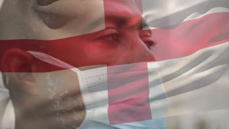 Animation-of-flag-of-england-waving-over-african-american-man-wearing-face-mask-in-city-street