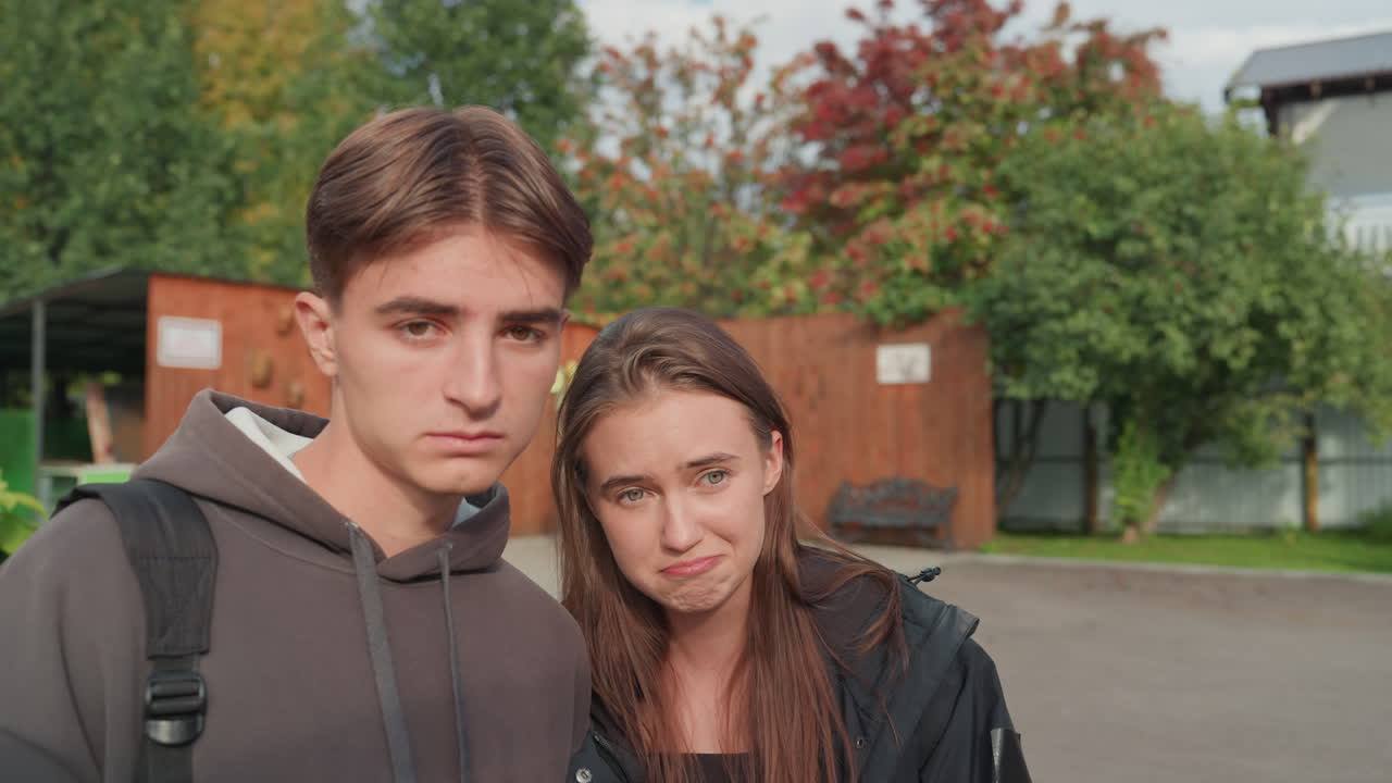 Portrett av hvite tenåringer på campusgårdsplassen under høsttrær, med ryggsekker og fritidsjakker, et intimt blikk og et forsiktig smil som formidler vennskap og forbindelse, mykt sollys og avslappet student