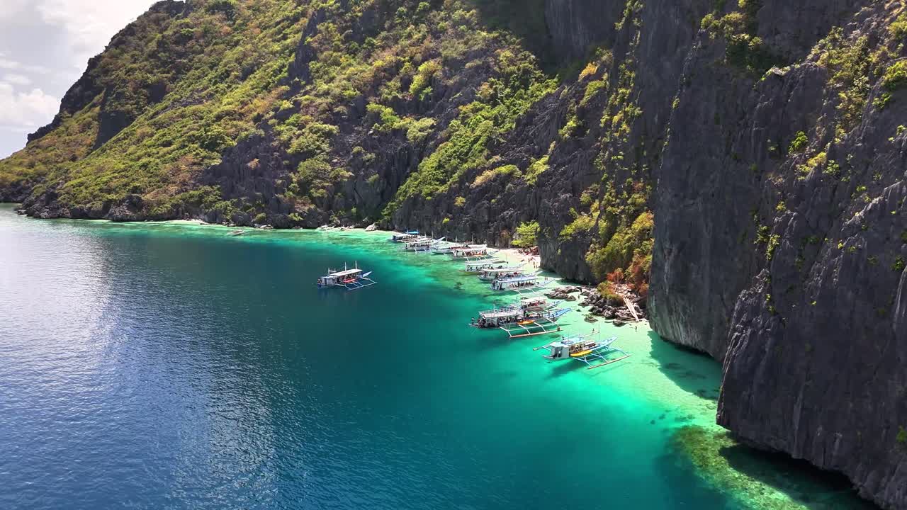 Traditional outrigger boats float on turquoise waters near limestone cliffs and lush forest at Talisay Beach on Tapiutan Island, Philippines, offering a serene tropical seascape and coastal beauty