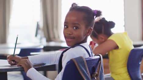 portrait of happy african american schoolgirl sitting at classroom, making notes, looking at camera