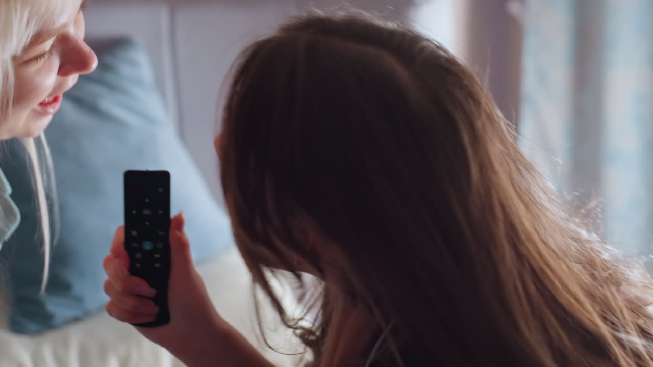 Close up of girl in black pajamas sitting on bed holding remote like microphone while dramatically singing with intense facial expression during lighthearted indoor moment filled with fun and energy