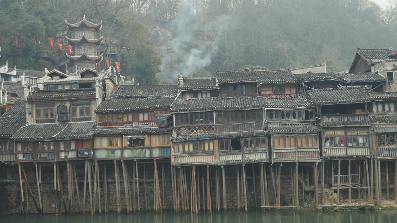 Casas tradicionais sobre palafitas ao longo do rio na Cidade Antiga de Fenghuang exibem arquitetura de madeira histórica e arredores tranquilos com fumaça subindo ao longe