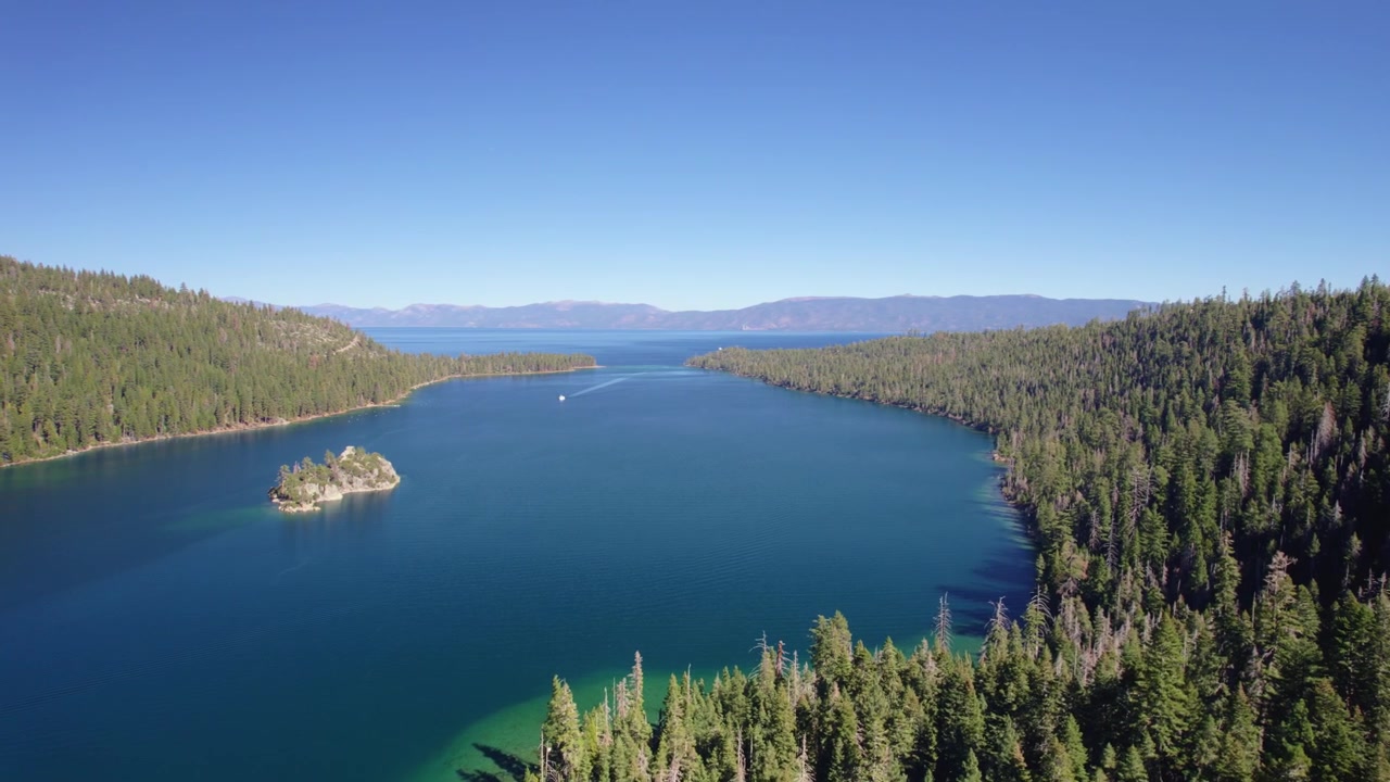 Un lento sorvolo cinematografico con drone sopra una fitta foresta alpina che rivela Emerald Bay e Fannette Island nel Lago Tahoe con acque di un blu intenso.