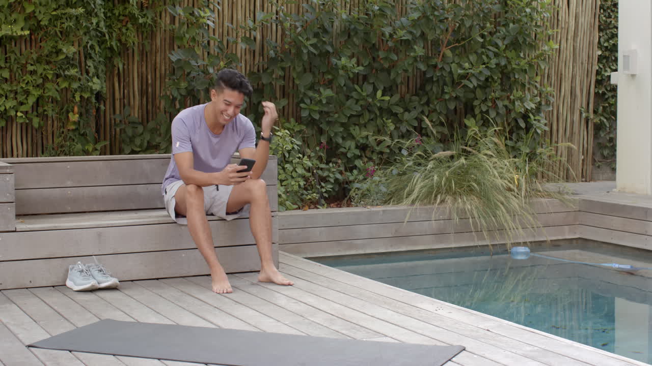 Sidder ved poolen, mand smiler og bruger smartphone, slapper af udendørs, med plads til tekst.