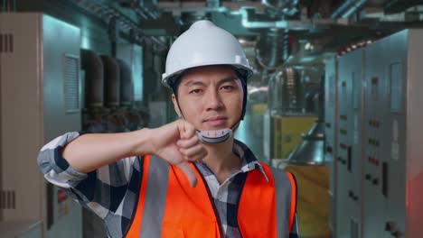 close up of asian male engineer with safety helmet showing thumbs down gesture and shaking his head while standing in engine control room, work of electrical generators