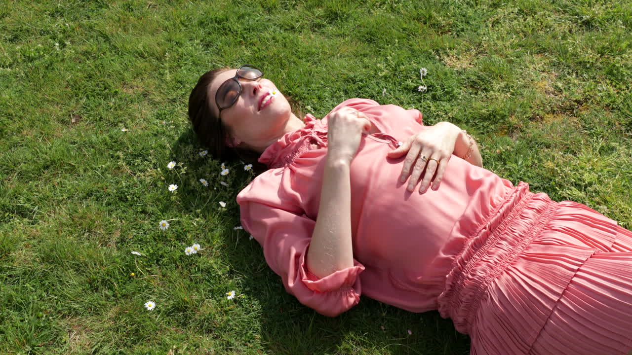 핑크색 드레스를 입은 아름다운 여성이 태양 아래 풀밭에서 휴식을 취하고 있습니다.