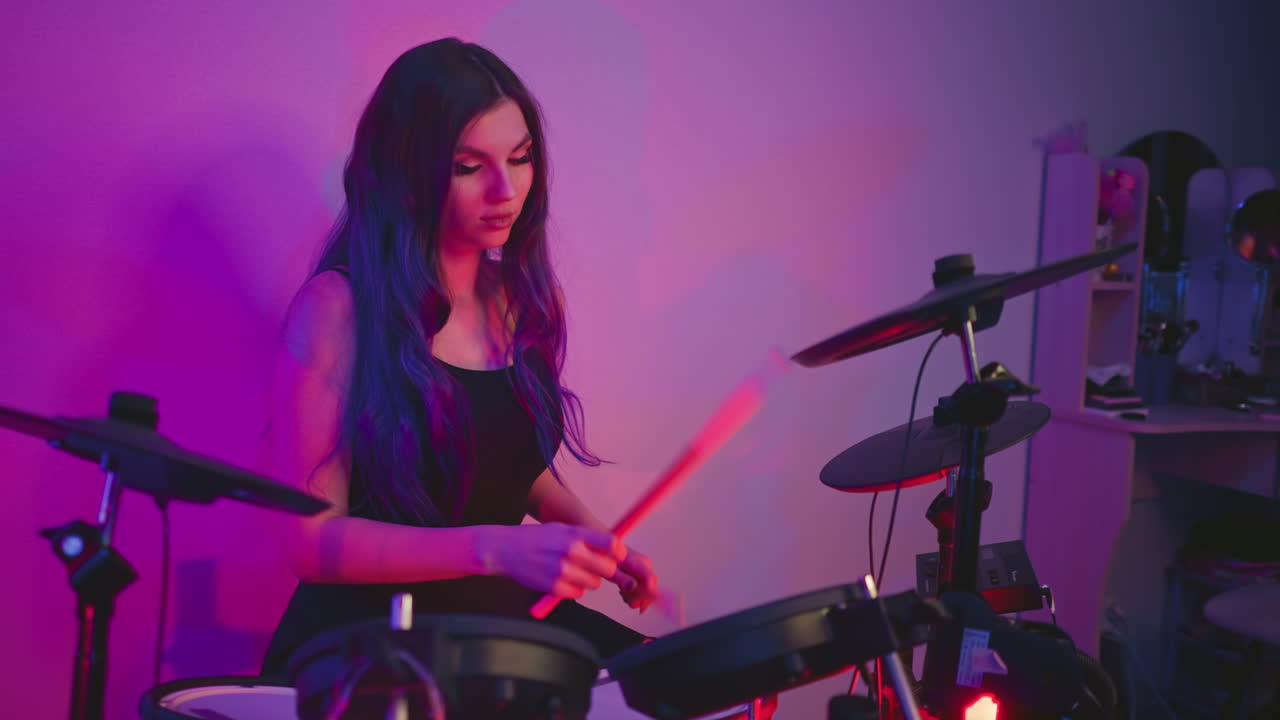 Close up of woman practicing drums on electronic kit in home studio lit with pink and purple neon lights creating ambient mood as she focuses on rhythm and beats during rehearsal session