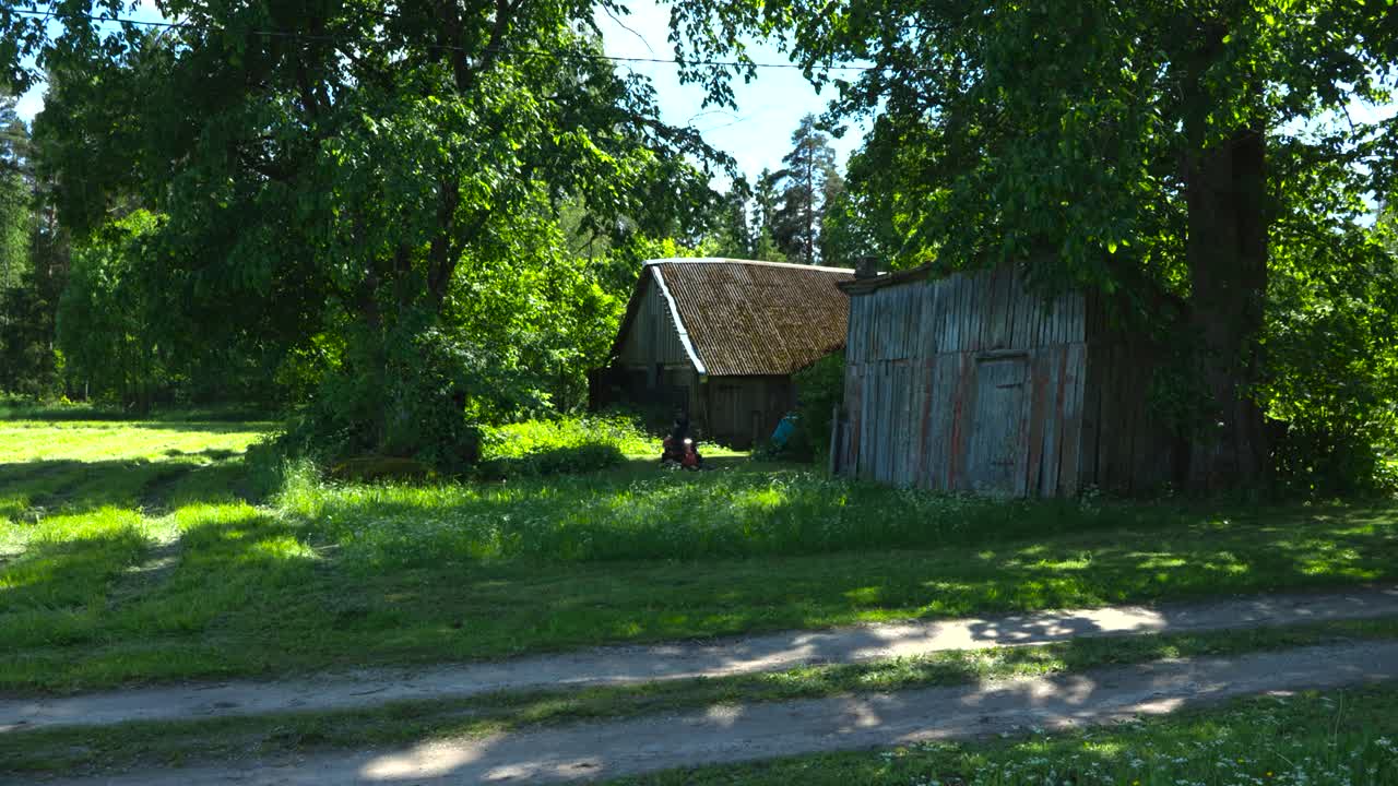 Gorgeous static footage of a man with black clothing and tattoos using a gasoline powered lawn mower to cut green grass in timelapse or time lapse mode at a sunny countryside garden, old barn visible.