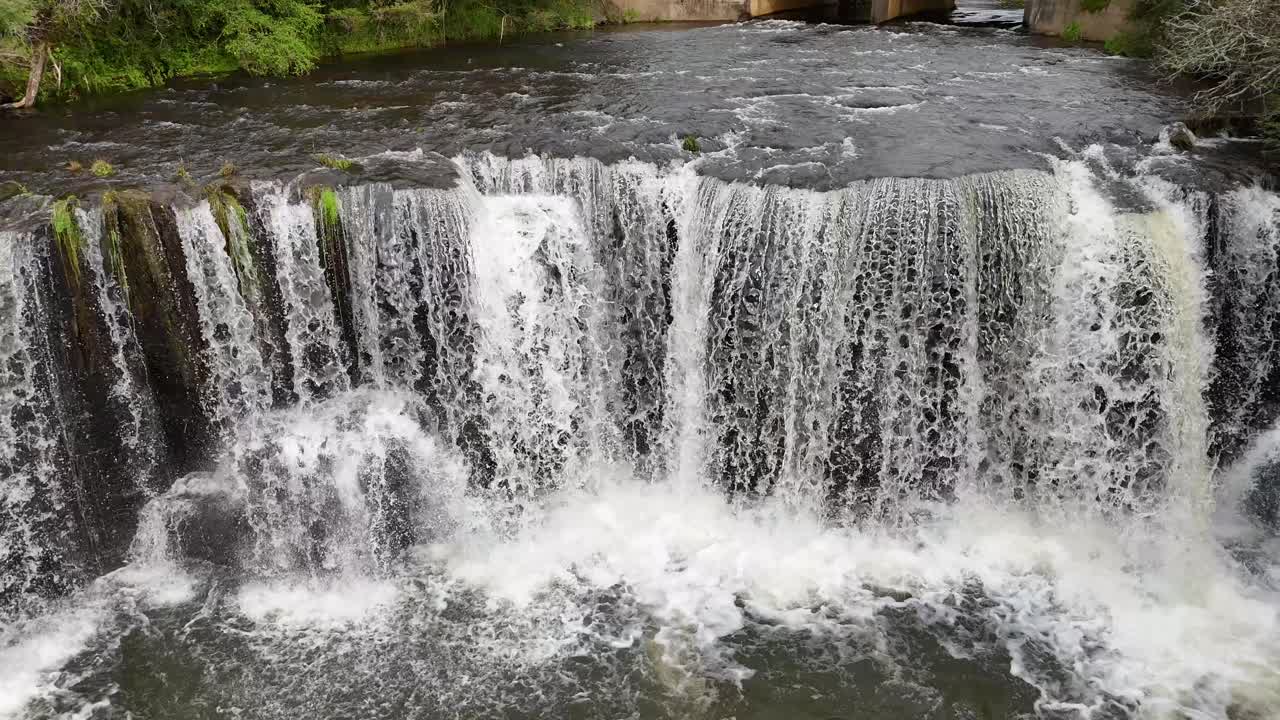 Drone camera descends above flowing river, revealing water cascading over concrete weir in daylight