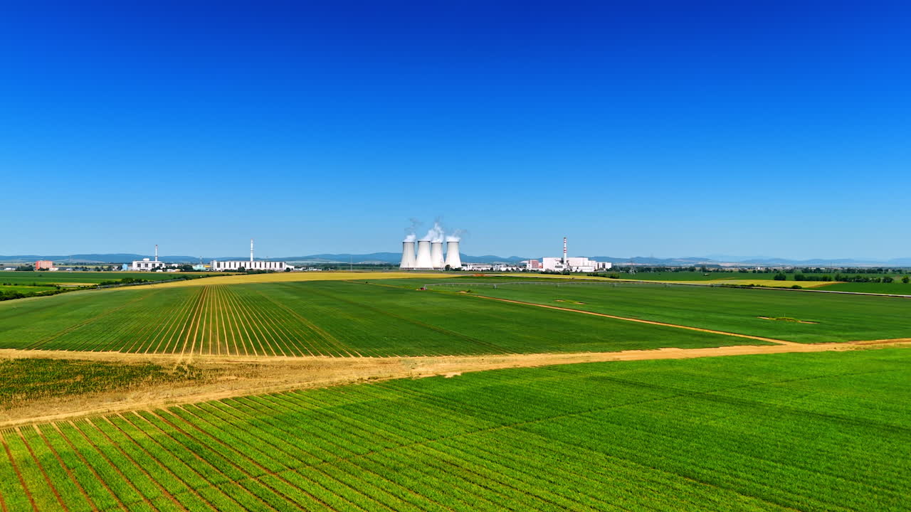 Fields by power plant, blue sky. Vast green fields stretch out towards a large industrial power plant set against a clear blue sky on a sunny day