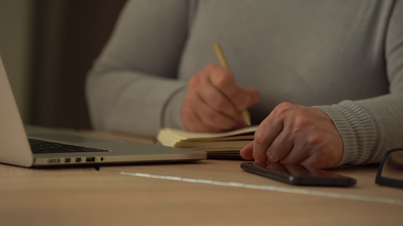 Close up wrinkled male hands writing information. Old mature woman working at office, using computer, handwriting notes