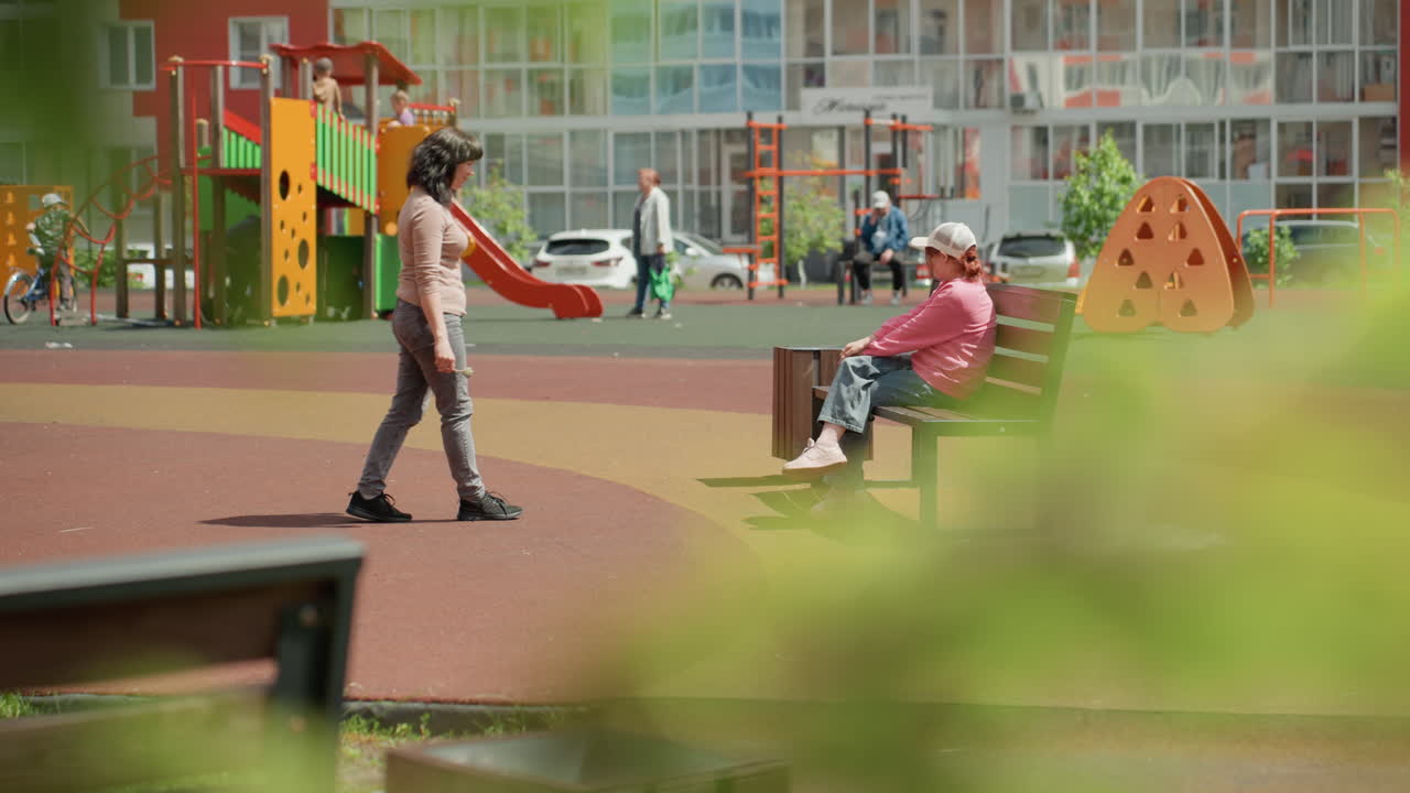 City Passerby Approaches Teenager Seated With Pet On Playground Bench, Bright Children Play Structures And Modern Housing In Distance, Moment Of Brief Conversation And Exchange, Casual Attire, Calm