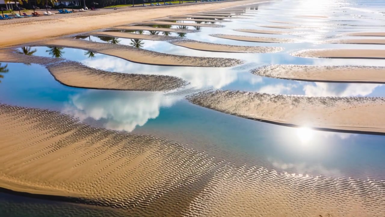Drone che plana su una piana di marea rivelando banchi di sabbia, palme, il bagliore del sole e impronte, mostrando motivi.