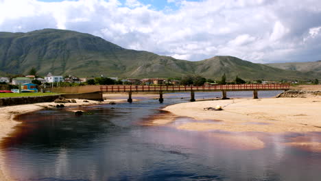 Kleinmond-lagoon-water-flowing-under-iconic-foot-bridge-toward-ocean,-riser-shot