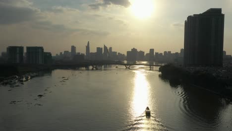 Seguimiento-De-Un-Barco-Fluvial-Tradicional-A-Lo-Largo-Del-Río-Saigon,-Vietnam-Desde-Un-Dron-Con-Vistas-Al-Horizonte-De-La-Ciudad-Moderna-Al-Atardecer