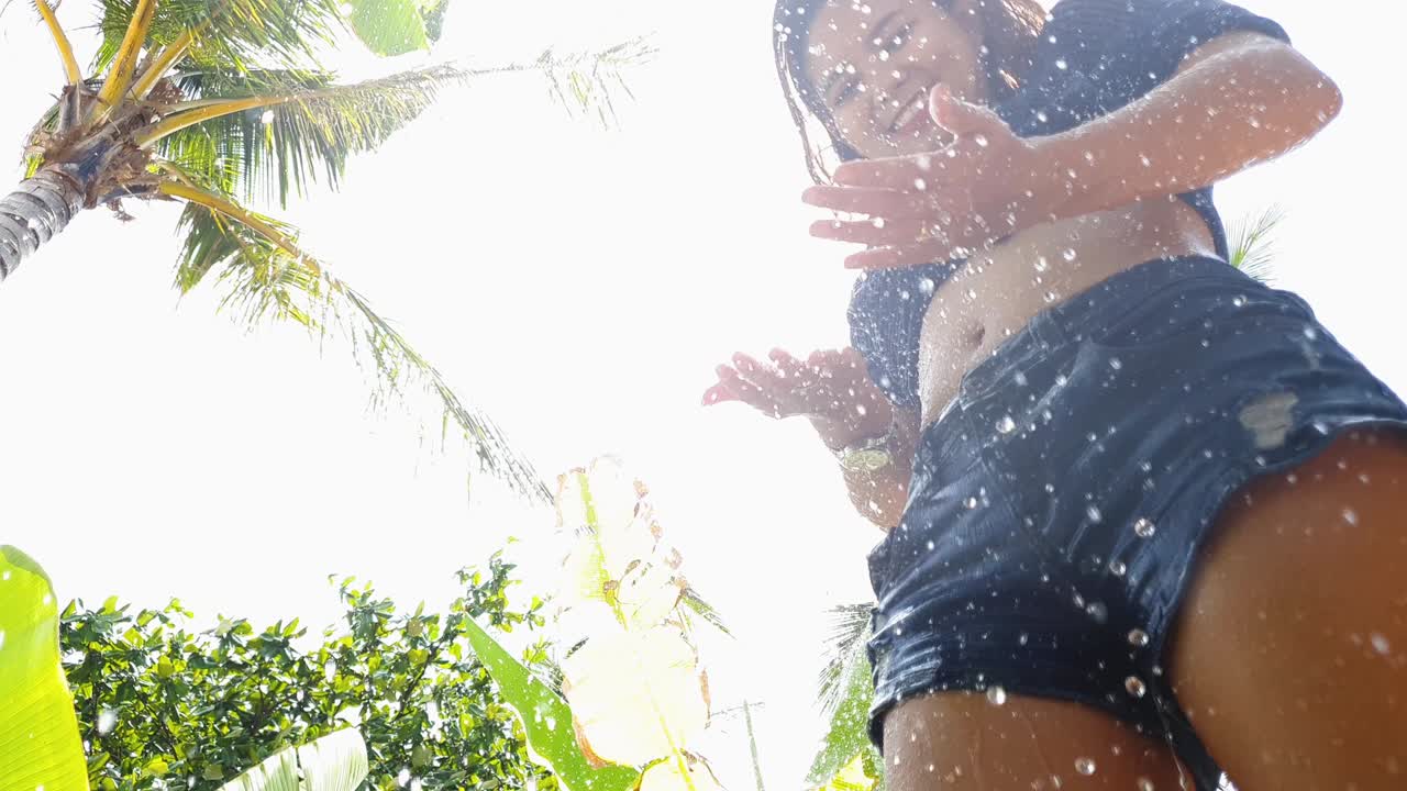 건강하고 튼튼한 다리와 몸매를 가진 활기찬 젊은 여성이 열대 해변에서 울창한 초록색으로 야외 샤워를 즐기고 있습니다.