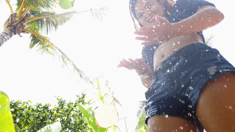 mirando a una joven animada con piernas sanas y un cuerpo en forma, disfrutando de una ducha al aire libre con exuberante vegetación en una playa tropical.