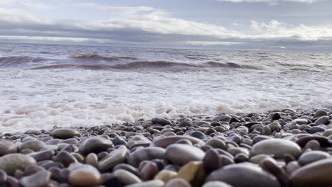 Seascape-of-the-waves-crashing-onto-a-pebble-beach-with-a-blue-sky-and-white-clouds