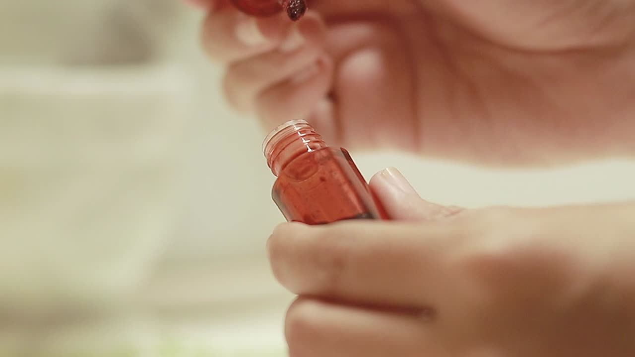 Hands holding and opening a tube of red lip tint as part of a makeup routine in a close-up shot against a beige background