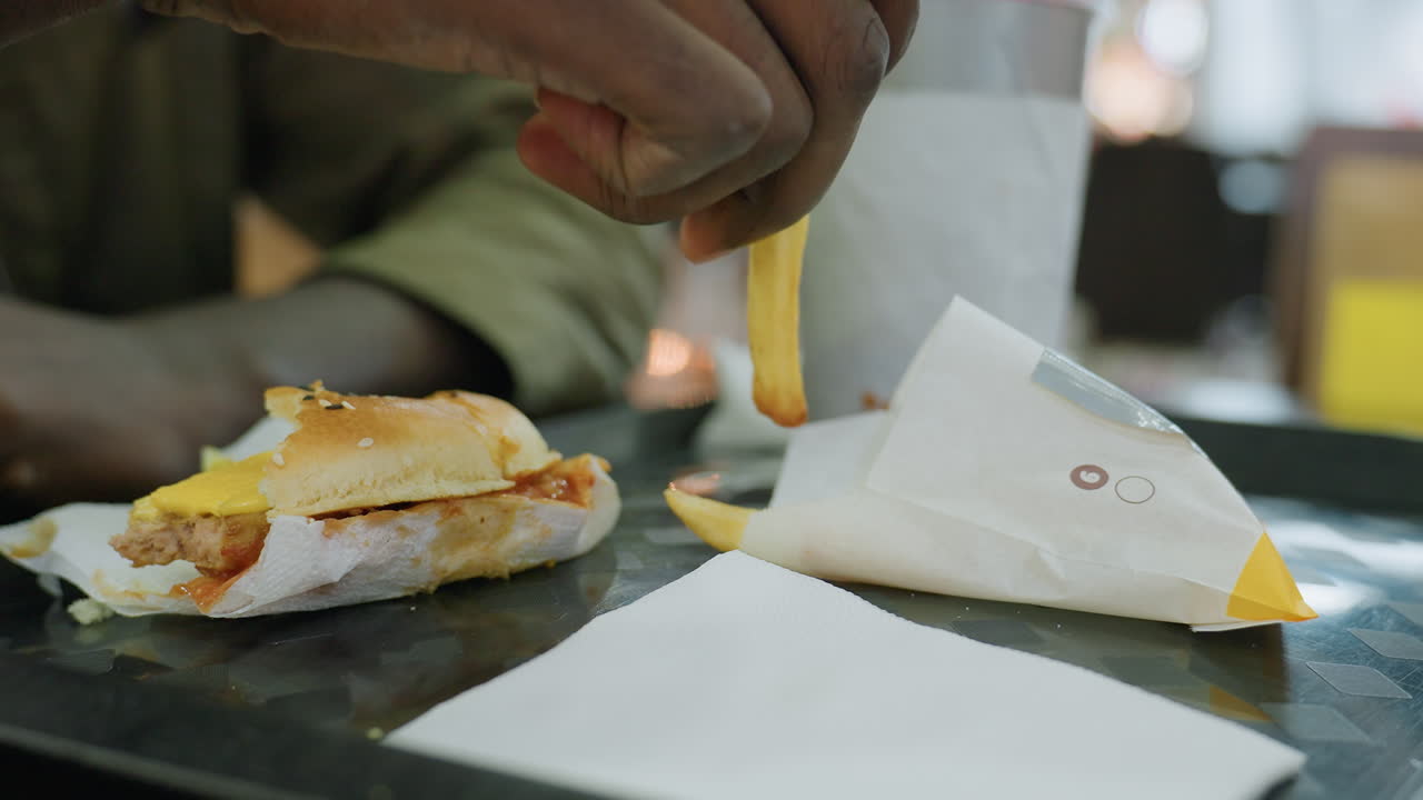Close up of meal on tray showing burger with cheese and fries in wrapper as partial view of person holding drink reaches to grab fries and eat indoors with blurred background