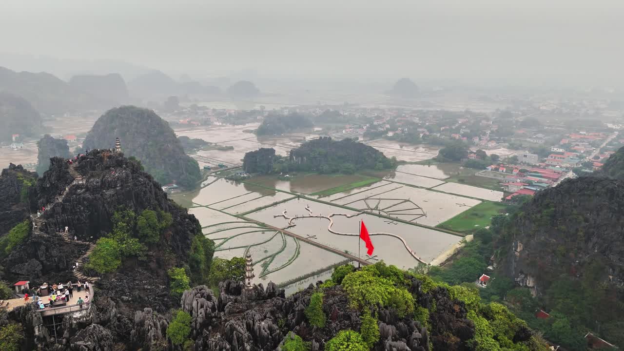 Panoramic aerial view of Dragon Hill in Tam Coc, Ninh Binh, Vietnam, with winding stone stairs, lush greenery, limestone peaks, and rice fields stretching into the misty background