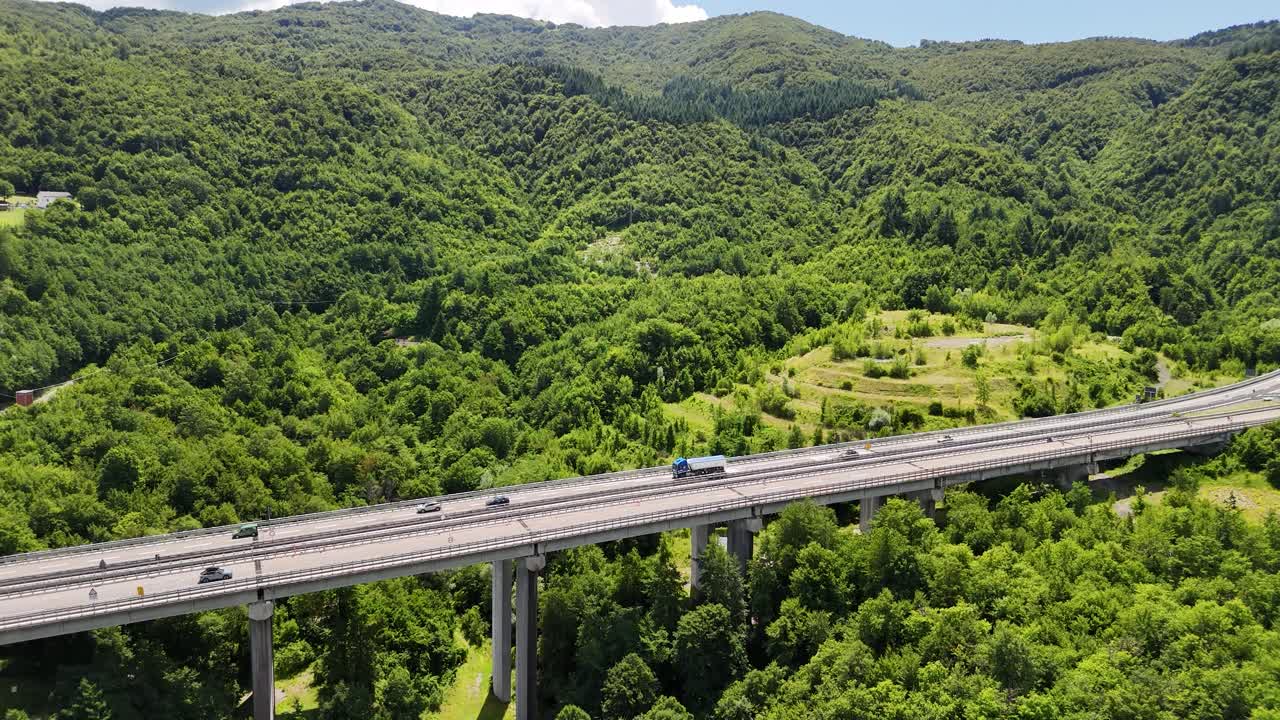 A red truck drives along a highway high above the ground, surrounded by vibrant green hills and valleys under a bright blue sky with fluffy clouds