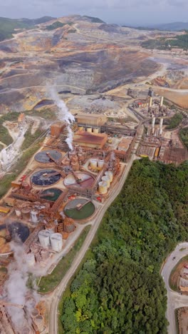 expansive backward aerial at barrick gold mine in cotuí, dominican republic, with industrial tanks, smokestacks, roads, and a vast open-pit mine. vertical shot