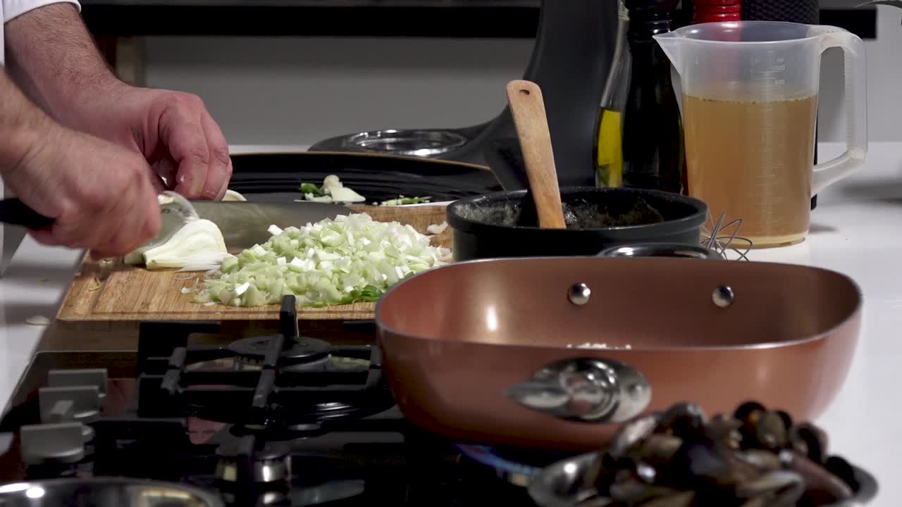 Skilled cook chops vegetables and prepares ingredients for a seafood pasta dish in a contemporary kitchen. Fresh clams, onions, and olive oil create a flavorful experience