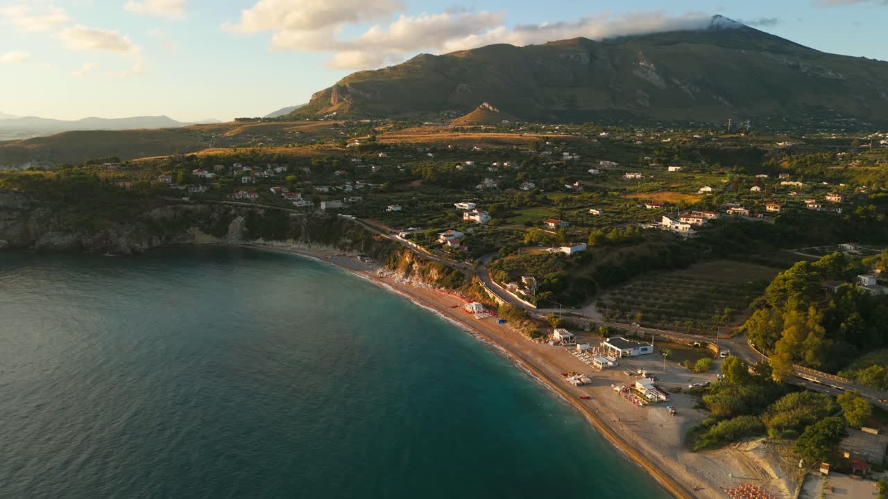 Orbiting aerial drone shot over empty Guidaloca Beach in Sicily, Italy, showing golden sunrise, waves, cliffs, and lush natural scenery