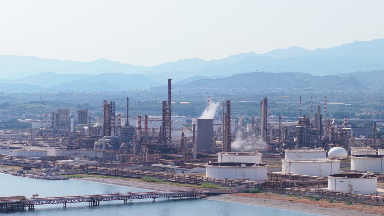 Drone wide shot of an oil refinery complex with storage tanks by the sea in Sicily, mountains in the background, and smoke rising from chimneys