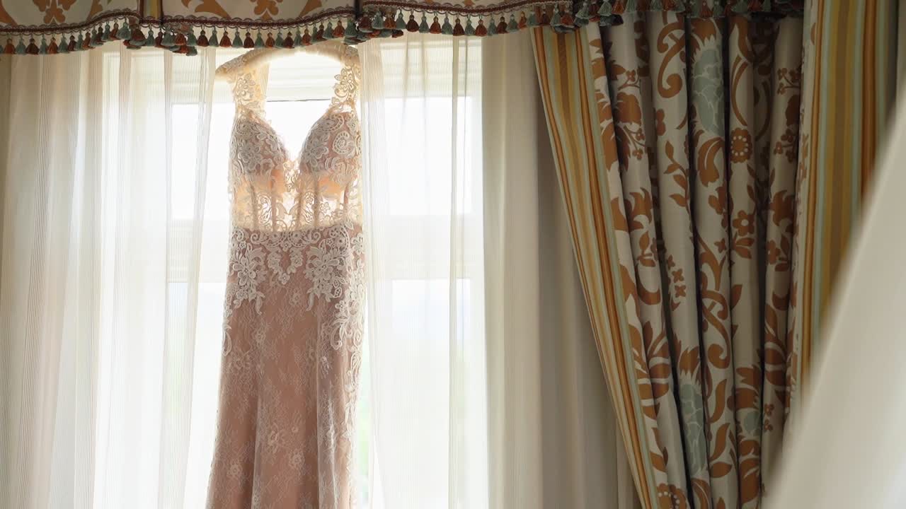 하얀 커튼과 장식용 커튼이 있는 밝은 창문에 매달려 있는 살색의 분홍색 웨딩드레스