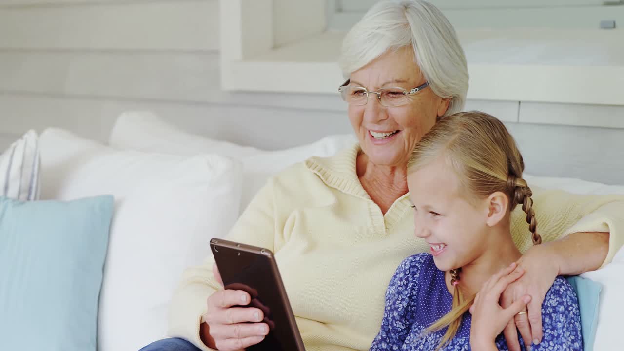 Happy grandmother and little girl seating on sofa and using tablet 4K 4k
