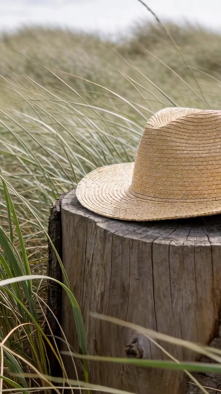 Video verticale: Panoramica della telecamera su un cappello di paglia intrecciato, rivelando i dettagli del cappello, con erbe delle dune che ondeggiano.