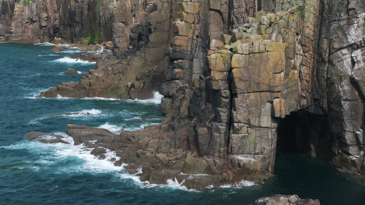 Drone circles coastal cave near Lands End with waves crashing against cliff walls, natural backdrop background as water meets rock